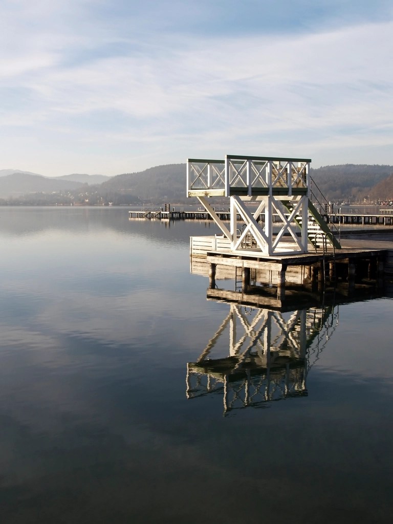 En brygga vid en lugn sjö med reflektioner i vattnet och omgivande berg i bakgrunden.